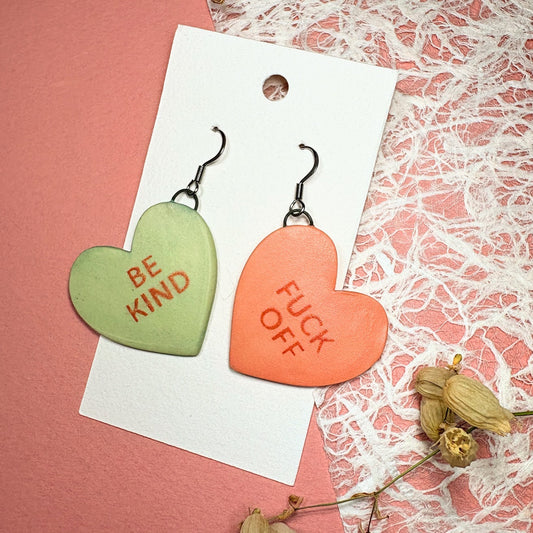 Heart-shaped earrings with 'BE KIND' and 'FUCK OFF' text on a pink background with lace.