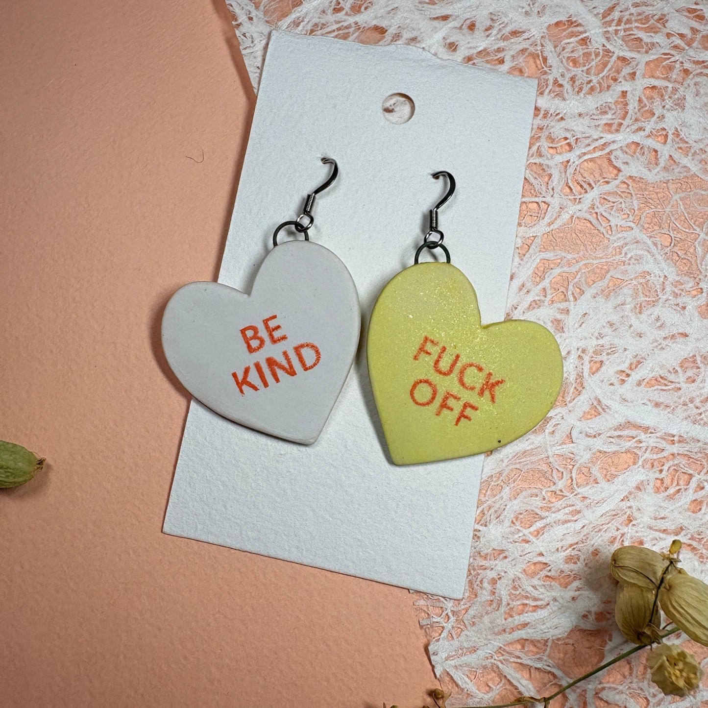 Heart-shaped earrings with 'FUCK OFF' and 'BE KIND' text on a textured surface.
